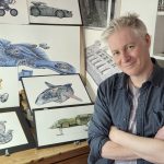 John R Mullaney with some of his art in his Reading studio. He has his arms crossed and he is looking at the camera
