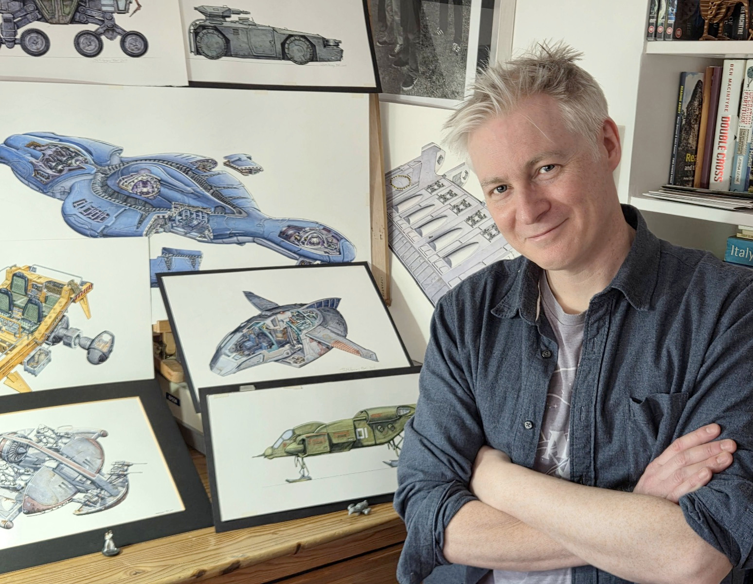 John R Mullaney with some of his art in his Reading studio. He has his arms crossed and he is looking at the camera
