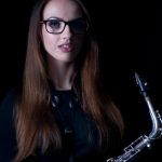 Alex Clarke holding a saxophone. The picture is dimly lit and the she is in half-shadow