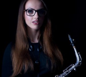Alex Clarke holding a saxophone. The picture is dimly lit and the she is in half-shadow