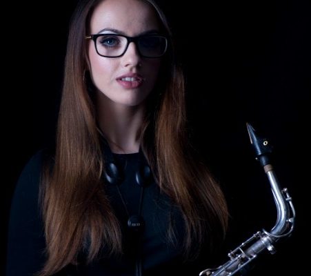 Alex Clarke holding a saxophone. The picture is dimly lit and the she is in half-shadow