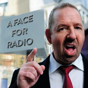 Alfie Moore standing next to a New Scotland Yard sign that has been tweaked to read 'A Face For Radio'. Alfie is pointing and has his mouth open as if in mid-rant.