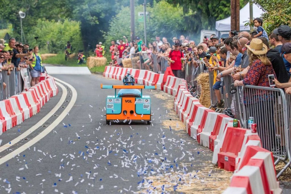 A racer in the Burghfield Box Kart Bash heads to the finish line with ticker tape on the ground and an audience watching behind a red and white barrier