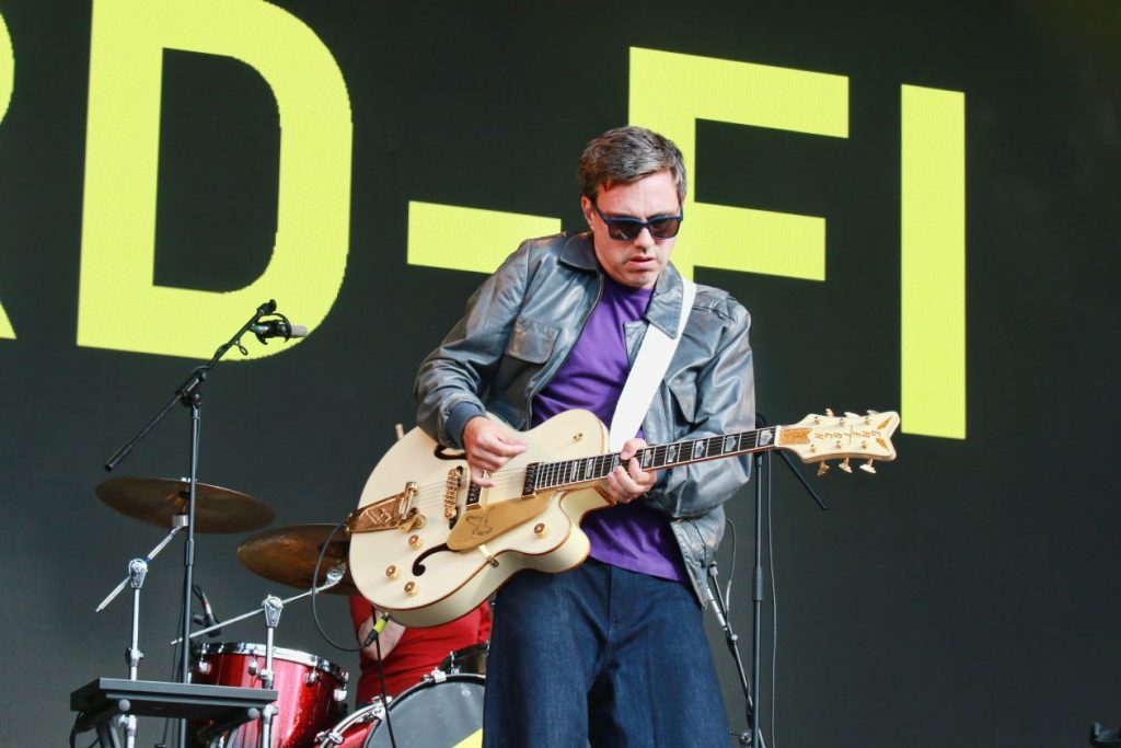 A man playing a guitar on a stage. There are letters visible behind him - D, F and I