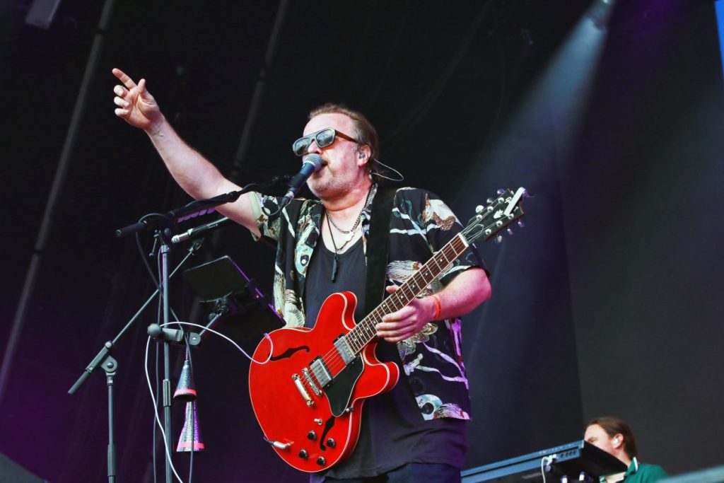 A man on stage holding a guitar, wearing sunglasses and pointing upwards towards the sky