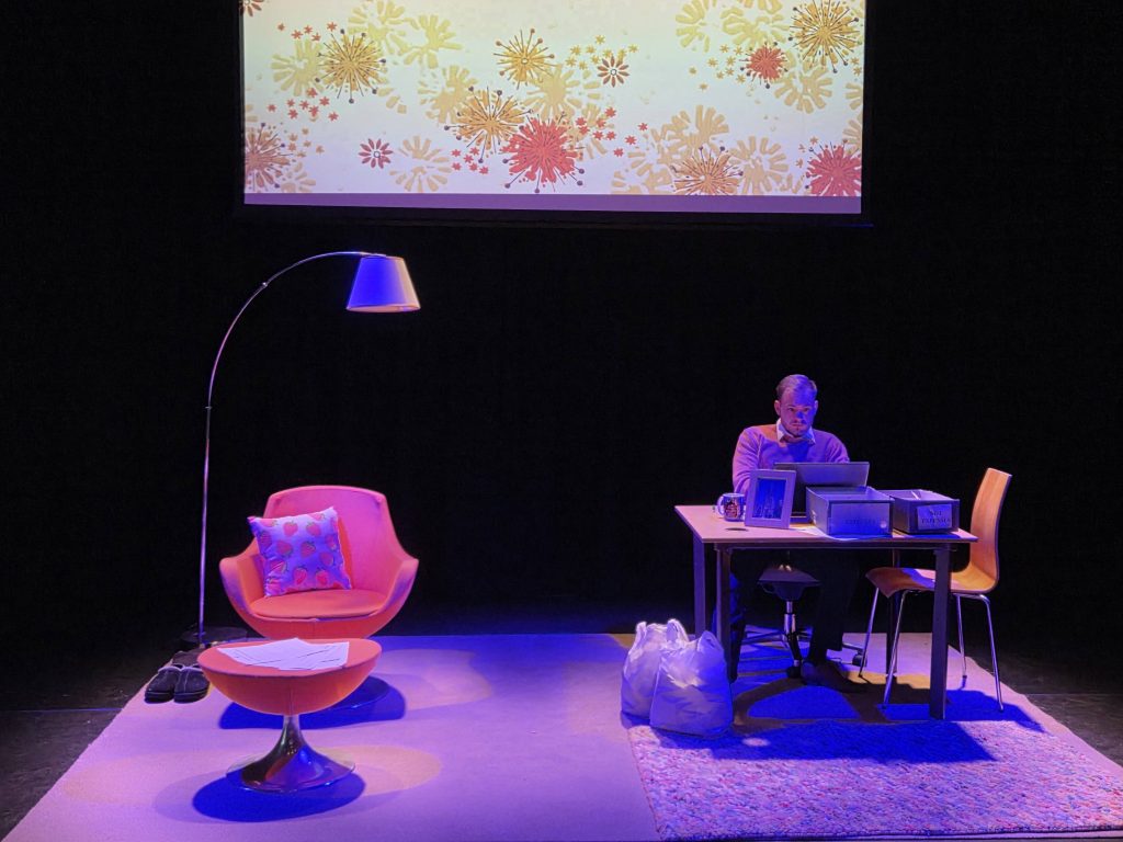 The sparse set for The Man includes a chair, a footstall, and a lamp on the left side, with a desk and chairs on the right. Ben Lewis is seated at the desk