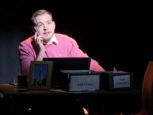 Ben Lewis in The Man. He is seated at a desk, holding a mobile phone. In front of him is a photo, and two boxes. One is labelled EXPENSES, the other NOT EXPENSES