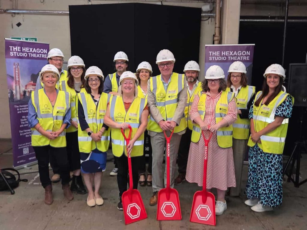 A group of people in hard hats and hi-vis vests. Three are holding red spades
