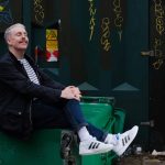 Carl Donnelly sitting back, cross-legged holding his knee with his hands. He has a moustache