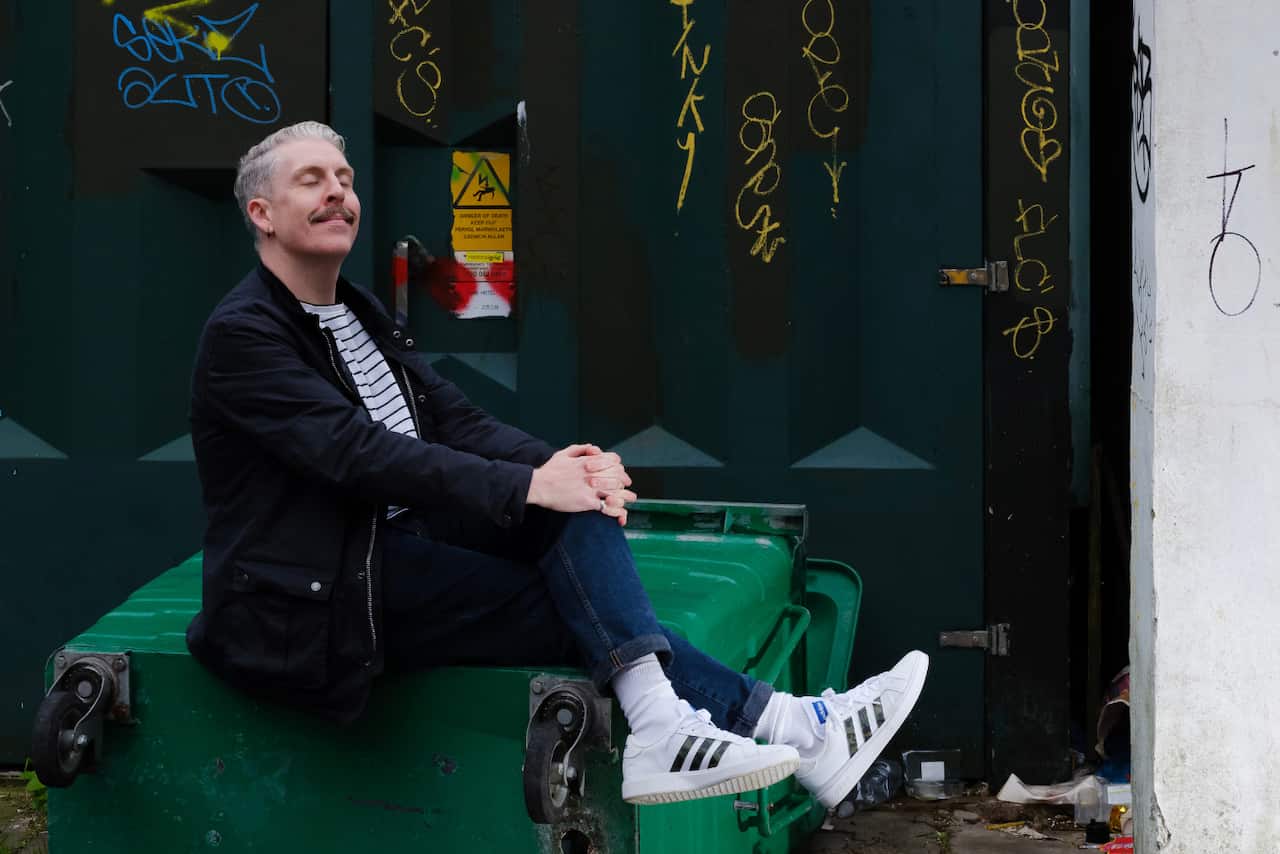 Carl Donnelly sitting back, cross-legged holding his knee with his hands. He has a moustache