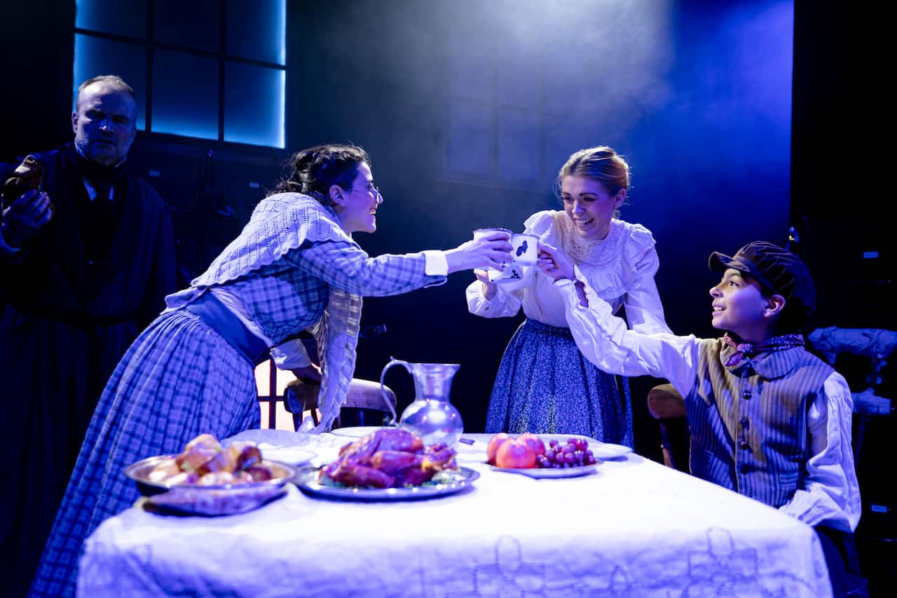 Three people around a table, the one seated is Tiny Tim