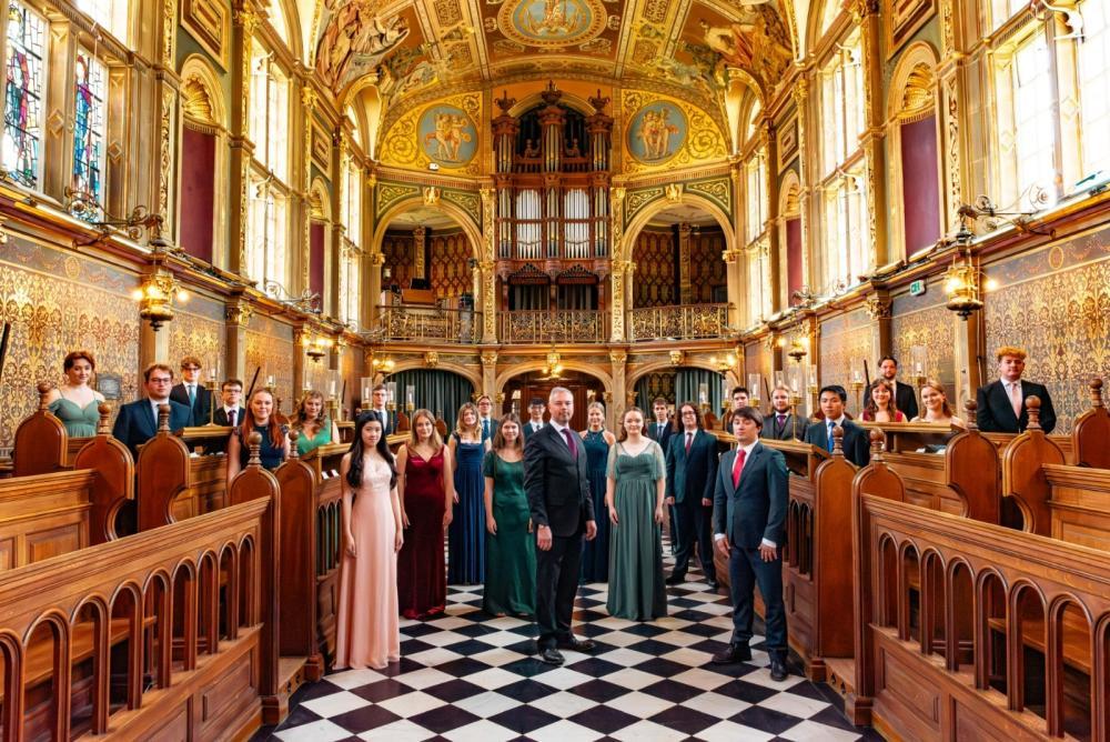 The Choir of Royal Holloway standing in a church. There is a black and white chequered floor, church pews and ornate walls