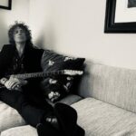 Kavus Torabi reclining on a sofa holding an instrument. The picture is in black and white