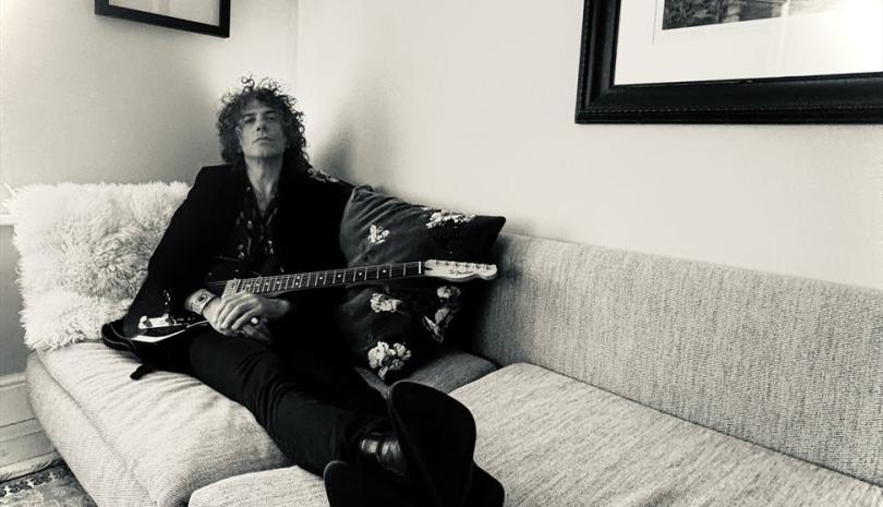 Kavus Torabi reclining on a sofa holding an instrument. The picture is in black and white