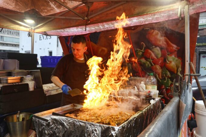 A man cooking at a grill, with flames coming out of it at Blue Collar Corner