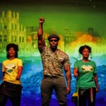 A vibrant stage scene from the musical Jamaica Love. Cast members are seen performing with energy, surrounded by a live band. The lighting is warm and evocative, capturing a moment of joyous musical celebration that represents the evolution of ska and reggae