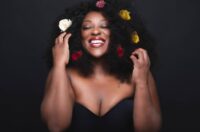 Judi Love with her eyes closed and arms bent and pointing upwards. She has a massive grin on her face and looks content