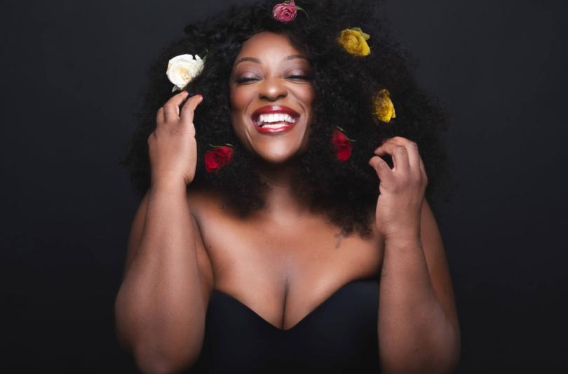 Judi Love with her eyes closed and arms bent and pointing upwards. She has a massive grin on her face and looks content