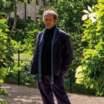 A portrait of gardener and broadcaster Monty Don. He is smiling warmly, dressed in his familiar outdoor workwear, set against a lush green garden background that reflects his lifelong connection to British horticulture