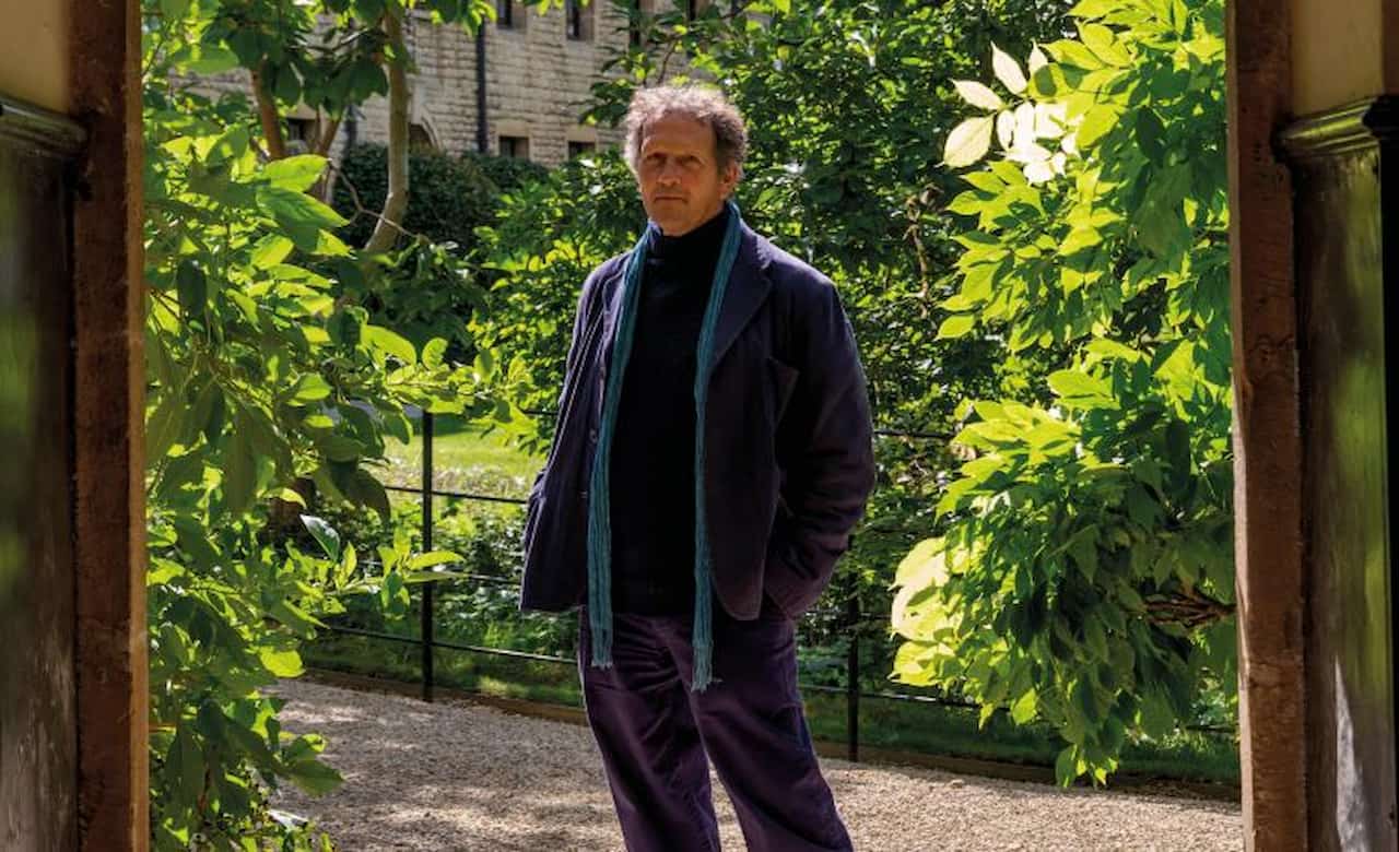A portrait of gardener and broadcaster Monty Don. He is smiling warmly, dressed in his familiar outdoor workwear, set against a lush green garden background that reflects his lifelong connection to British horticulture