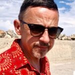 A close-up outdoor portrait shot of Peter Harris, known as Blu Peter, looking slightly downward and to the right of the frame. He is wearing black-framed sunglasses, a red short-sleeved shirt patterned with gold geometric and circular designs, and has short dark hair and a trimmed goatee. The background shows an outdoor setting with a light-coloured stone or concrete wall under a blue sky with scattered white clouds.