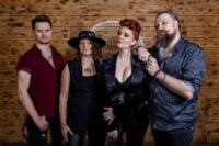 A studio portrait of the four members of the rock band Brave Rival standing side by side against a horizontal wooden panel background. From left to right: a man with short dark hair wearing a dark red short-sleeved patterned shirt and a silver necklace; a woman with wavy brown hair wearing a black wide-brimmed hat, a black sleeveless top, and a large round silver pendant necklace; a woman with styled red hair pulled up, wearing a black satin-style blazer with silver embellishments on the shoulders over a black lace top; and a man with a prominent beard and moustache wearing a dark paisley patterned button-up shirt with his left hand resting on the shoulder of the woman next to him.