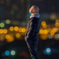 A full-length profile photograph of John McCusker standing and looking upwards and to the left with his eyes closed. He is wearing a dark, quilted puffer jacket over a grey hooded sweatshirt with the hood down, paired with dark jeans. His hands are tucked into his jacket pockets. The background is completely out of focus, showing a dark night scene illuminated by blurry golden, green, and blue bokeh light circles from a city or traffic.