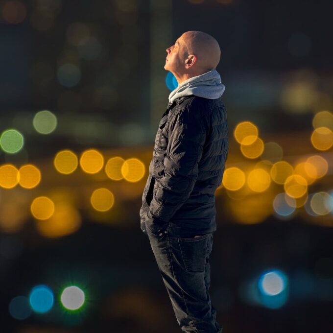 A full-length profile photograph of John McCusker standing and looking upwards and to the left with his eyes closed. He is wearing a dark, quilted puffer jacket over a grey hooded sweatshirt with the hood down, paired with dark jeans. His hands are tucked into his jacket pockets. The background is completely out of focus, showing a dark night scene illuminated by blurry golden, green, and blue bokeh light circles from a city or traffic.