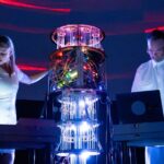 A live performance photograph of the audiovisual duo Movement81 on a dark stage illuminated by intense blue and white lights. In the centre stands a tall, cylindrical piece of electronic equipment or a custom modular synthesizer rig, glowing with internal green, yellow, and red lights and featuring hanging clear tubes or wires that reflect bright blue and white light at the base. On the left, a woman with blonde hair tied back wears a fitted white short-sleeved dress and stands behind a electronic keyboard or console, reaching out one hand toward the central equipment. On the right, a man wearing glasses and a white button-up short-sleeved shirt stands looking down at a laptop computer and a mixing console. The background features dark curved walls with faint horizontal red light strips overhead.