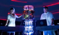 A live performance photograph of the audiovisual duo Movement81 on a dark stage illuminated by intense blue and white lights. In the centre stands a tall, cylindrical piece of electronic equipment or a custom modular synthesizer rig, glowing with internal green, yellow, and red lights and featuring hanging clear tubes or wires that reflect bright blue and white light at the base. On the left, a woman with blonde hair tied back wears a fitted white short-sleeved dress and stands behind a electronic keyboard or console, reaching out one hand toward the central equipment. On the right, a man wearing glasses and a white button-up short-sleeved shirt stands looking down at a laptop computer and a mixing console. The background features dark curved walls with faint horizontal red light strips overhead.