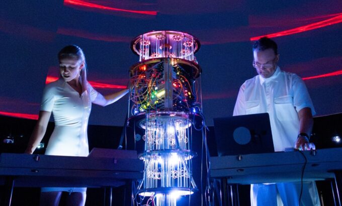 A live performance photograph of the audiovisual duo Movement81 on a dark stage illuminated by intense blue and white lights. In the centre stands a tall, cylindrical piece of electronic equipment or a custom modular synthesizer rig, glowing with internal green, yellow, and red lights and featuring hanging clear tubes or wires that reflect bright blue and white light at the base. On the left, a woman with blonde hair tied back wears a fitted white short-sleeved dress and stands behind a electronic keyboard or console, reaching out one hand toward the central equipment. On the right, a man wearing glasses and a white button-up short-sleeved shirt stands looking down at a laptop computer and a mixing console. The background features dark curved walls with faint horizontal red light strips overhead.
