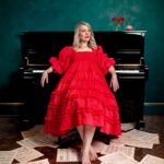 A studio portrait of comedian Rachel Parris sitting on a stool in front of a glossy black upright piano against a textured dark green wall. She is looking to the right with a slight smile and is wearing a bright red, tiered mid-length dress with puff sleeves and smocking across the bodice. She is barefoot with red nail polish on her toes, and her feet rest on loose sheets of musical manuscript paper scattered across a dark wooden parquet floor. On the top right of the piano sits a small clear glass vase containing three red roses, next to a small red novelty wind-up chattering teeth toy.