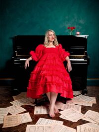 A studio portrait of comedian Rachel Parris sitting on a stool in front of a glossy black upright piano against a textured dark green wall. She is looking to the right with a slight smile and is wearing a bright red, tiered mid-length dress with puff sleeves and smocking across the bodice. She is barefoot with red nail polish on her toes, and her feet rest on loose sheets of musical manuscript paper scattered across a dark wooden parquet floor. On the top right of the piano sits a small clear glass vase containing three red roses, next to a small red novelty wind-up chattering teeth toy.