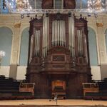 The Father Willis pipe organ inside Reading Town Hall, featuring a large, dark wooden casing with silver pipes, viewed from the front of the stage. The grand organ stands centrally against a light blue wall with decorative arches and gold accents, flanked by tiered seating and modern stage lighting rigs overhead.
