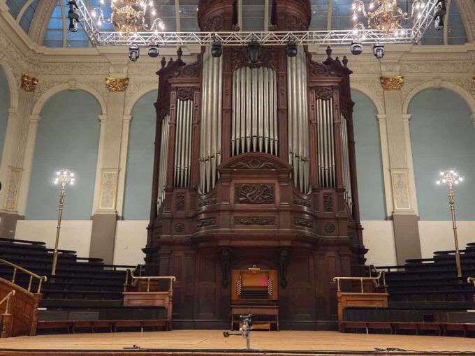The Father Willis pipe organ inside Reading Town Hall, featuring a large, dark wooden casing with silver pipes, viewed from the front of the stage. The grand organ stands centrally against a light blue wall with decorative arches and gold accents, flanked by tiered seating and modern stage lighting rigs overhead.