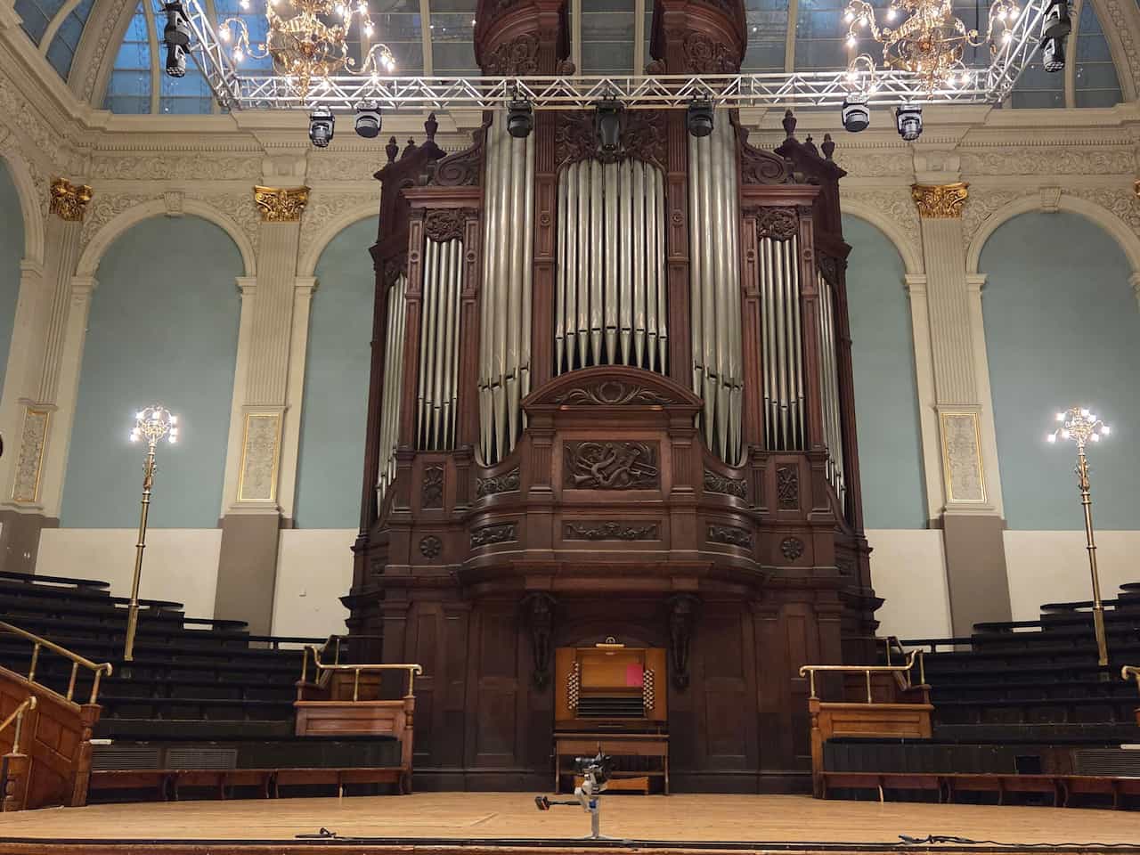 The Father Willis pipe organ inside Reading Town Hall, featuring a large, dark wooden casing with silver pipes, viewed from the front of the stage. The grand organ stands centrally against a light blue wall with decorative arches and gold accents, flanked by tiered seating and modern stage lighting rigs overhead.