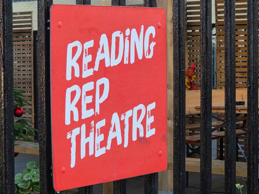 A close-up shot of a red square sign with the words Reading Rep Theatre painted on it in white hand-brushed lettering. The sign is attached to a black metal fence, with an outdoor seating area and wooden cladding visible in the blurred background.