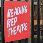 A close-up shot of a red square sign with the words Reading Rep Theatre painted on it in white hand-brushed lettering. The sign is attached to a black metal fence, with an outdoor seating area and wooden cladding visible in the blurred background.