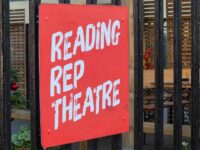 A close-up shot of a red square sign with the words Reading Rep Theatre painted on it in white hand-brushed lettering. The sign is attached to a black metal fence, with an outdoor seating area and wooden cladding visible in the blurred background.