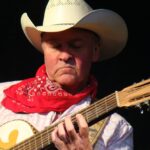 A close-up of Paul Young holding a guitar and wearing a hat