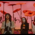An image of the alternative rock band Black Bordello standing in a surreal, dreamlike field under a deep pink and red sky. The background is dominated by oversized, giant red gerbera daisy-like flowers towering over the band. In the foreground, four band members are lined up, dressed in theatrical, gothic, and vaudeville-inspired attire featuring corsets, lace, and dark clothing. Two members have striking white face paint and dramatic dark makeup, while the others wear stylized clown-like cheek makeup. They hold single yellow sunflowers and look directly forward with serious, enigmatic expressions.