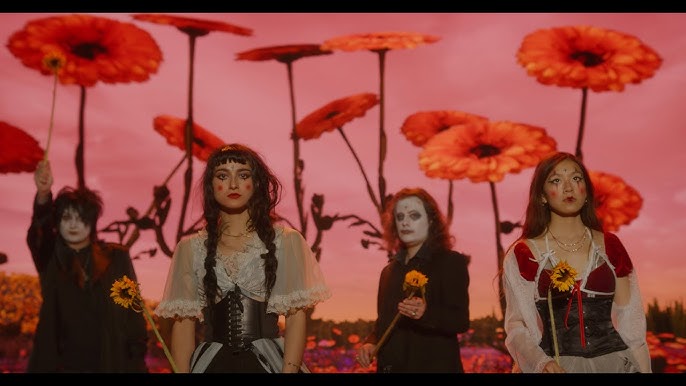 An image of the alternative rock band Black Bordello standing in a surreal, dreamlike field under a deep pink and red sky. The background is dominated by oversized, giant red gerbera daisy-like flowers towering over the band. In the foreground, four band members are lined up, dressed in theatrical, gothic, and vaudeville-inspired attire featuring corsets, lace, and dark clothing. Two members have striking white face paint and dramatic dark makeup, while the others wear stylized clown-like cheek makeup. They hold single yellow sunflowers and look directly forward with serious, enigmatic expressions.