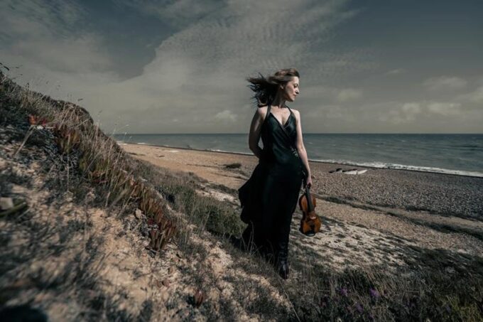 Violinist Fenella Humphreys stands on a grassy coastal hill overlooking a pebble beach and the sea under a dramatic, cloudy sky. She is wearing a long black gown, her hair is catching the wind, and she holds her violin by her side.
