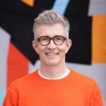 Headshot of Gareth Malone smiling at the camera, wearing an orange jumper over a white t-shirt and round tortoise-shell glasses, with a blurred colorful abstract mural in the background.