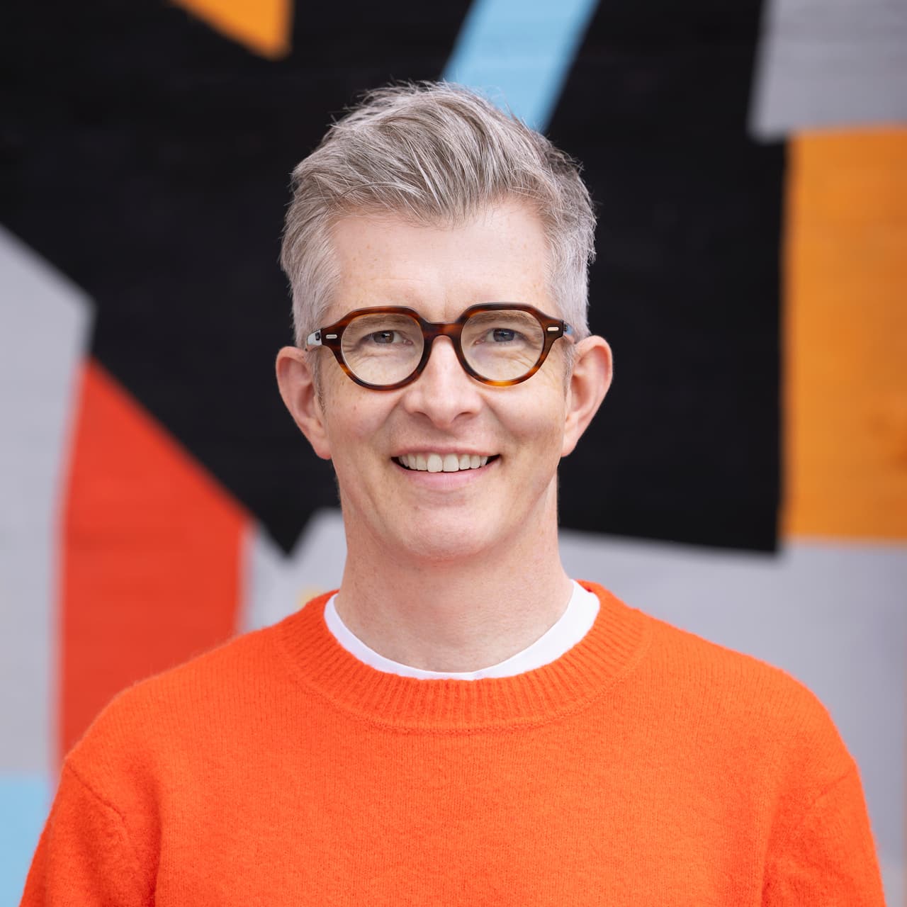 Headshot of Gareth Malone smiling at the camera, wearing an orange jumper over a white t-shirt and round tortoise-shell glasses, with a blurred colorful abstract mural in the background.