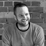 A black and white portrait of Paul Stacey smiling and looking to the right. He has short hair, a beard, and his arms are crossed while wearing a sweatshirt with a circular graphic of a bear. He is sitting in front of an exposed brick wall.