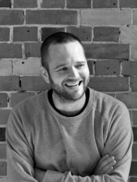 A black and white portrait of Paul Stacey smiling and looking to the right. He has short hair, a beard, and his arms are crossed while wearing a sweatshirt with a circular graphic of a bear. He is sitting in front of an exposed brick wall.