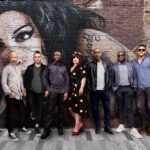 The eight members of The Amy Winehouse Band stand in a line on a paved street, posing in front of a brick wall featuring a large painted mural of Amy Winehouse. The musicians are dressed in smart-casual autumn attire, with a female vocalist in a black and white polka dot dress standing near the centre.