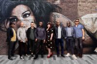 The eight members of The Amy Winehouse Band stand in a line on a paved street, posing in front of a brick wall featuring a large painted mural of Amy Winehouse. The musicians are dressed in smart-casual autumn attire, with a female vocalist in a black and white polka dot dress standing near the centre.
