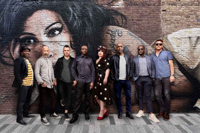 The eight members of The Amy Winehouse Band stand in a line on a paved street, posing in front of a brick wall featuring a large painted mural of Amy Winehouse. The musicians are dressed in smart-casual autumn attire, with a female vocalist in a black and white polka dot dress standing near the centre.